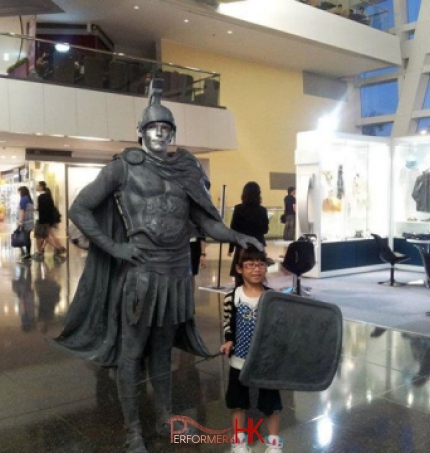 Human statue performer in silver centurion costume putting his hand on small girls head whilst posing 