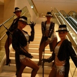 Four dancers in silver costumes, with black sequined coats and cane posing just before performing the Charleston dance.