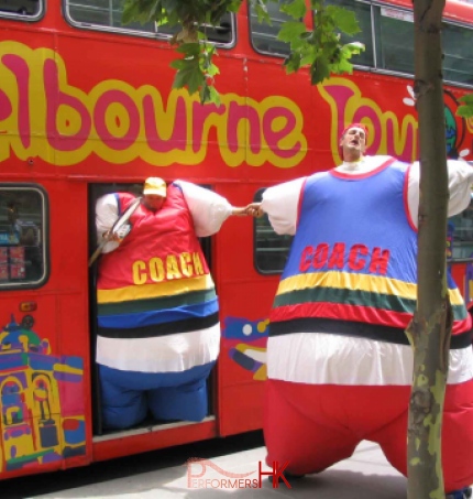 Hong Kong stilt-walker in a blue inflatable coach costume pulling other stilt-walker in a red inflatable coach costume from a bus