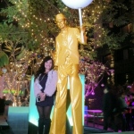 Golden stilt walker holding huge chupa chup and posing for picture with a lady