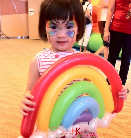 A little girl with butterfly face paint holding a rainbow balloon from a roving balloons clown at a fun day event