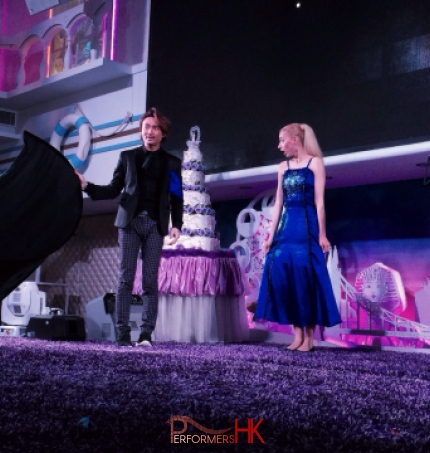 Magician in Hong Kong standing in front of a wedding cake ,holding a black cloth ,performing stage magic with is assistant who wearing a blue dress at a corporate event