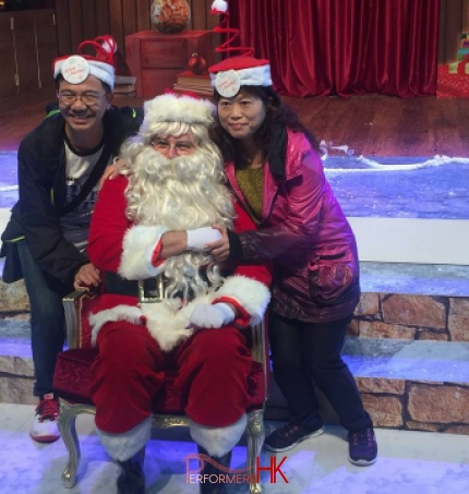 HK performer dress as Santa, taking picture with two tourists who wearing Xmas hat in front of the Xmas backdrop at a Hong Kong Christmas function.