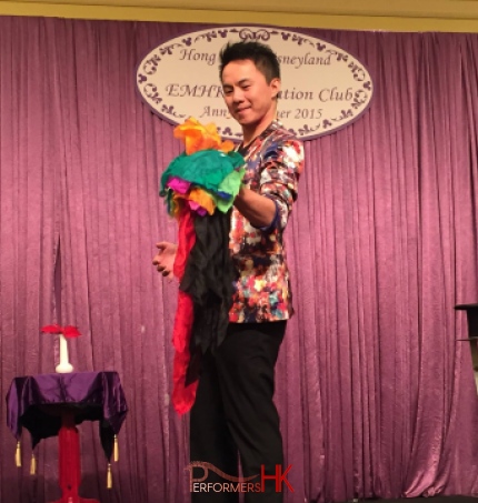 Magician in HK standing in the middle of magic hat and vase performing stage magic with colorful silks .