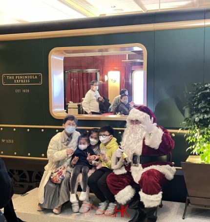 Santa performer in front of a train set up on a bench taking photos with 3 children and a mother in hong kong santa in deep red color costume 