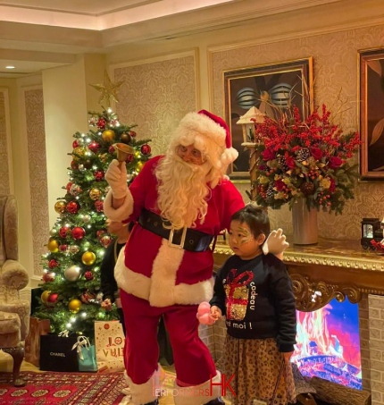 Santa Gerald standing in front of a christmas tree with 2 kids during Christmas time, at the penninsula hotel suite. Christmas tree is lit up 2025 Christmas Hong Kong, there is a faux fire place in the background